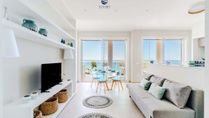 En moderne stue med TV, sofa og spiseplads med stole, der fører til en balkon med udsigt.