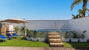 Un patio con césped artificial, una piscina, sillas de descanso y plantas en macetas.