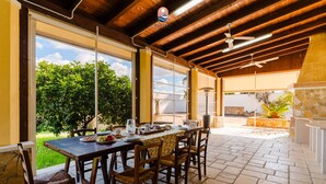 Un espacio al aire libre con comedor, mesa y sillas de madera, chimenea de piedra y vista a follaje.