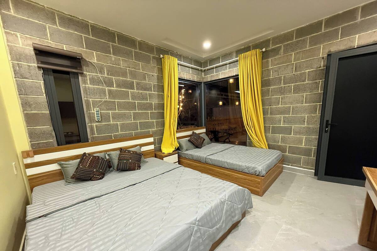 A bedroom with two beds, a brick wall, a window with yellow curtains, and a door.