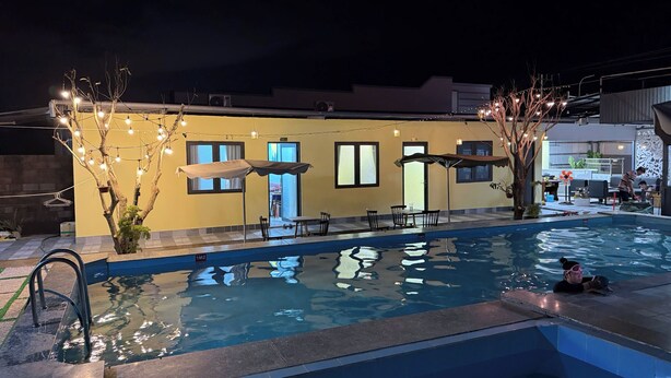 Una piscina con bordo piastrellato, un piccolo albero illuminato e un edificio sullo sfondo con finestre e porte.