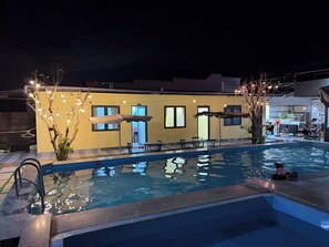 Una piscina con borde de baldosas, un pequeño árbol con luces y un edificio con ventanas y puertas al fondo.