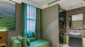 Ein modernes Zimmer mit einem grünen Sofa, einem großen Fenster mit Vorhängen, einem Badezimmer mit Waschbecken und Spiegel sowie einem Wandbild einer Landschaft.