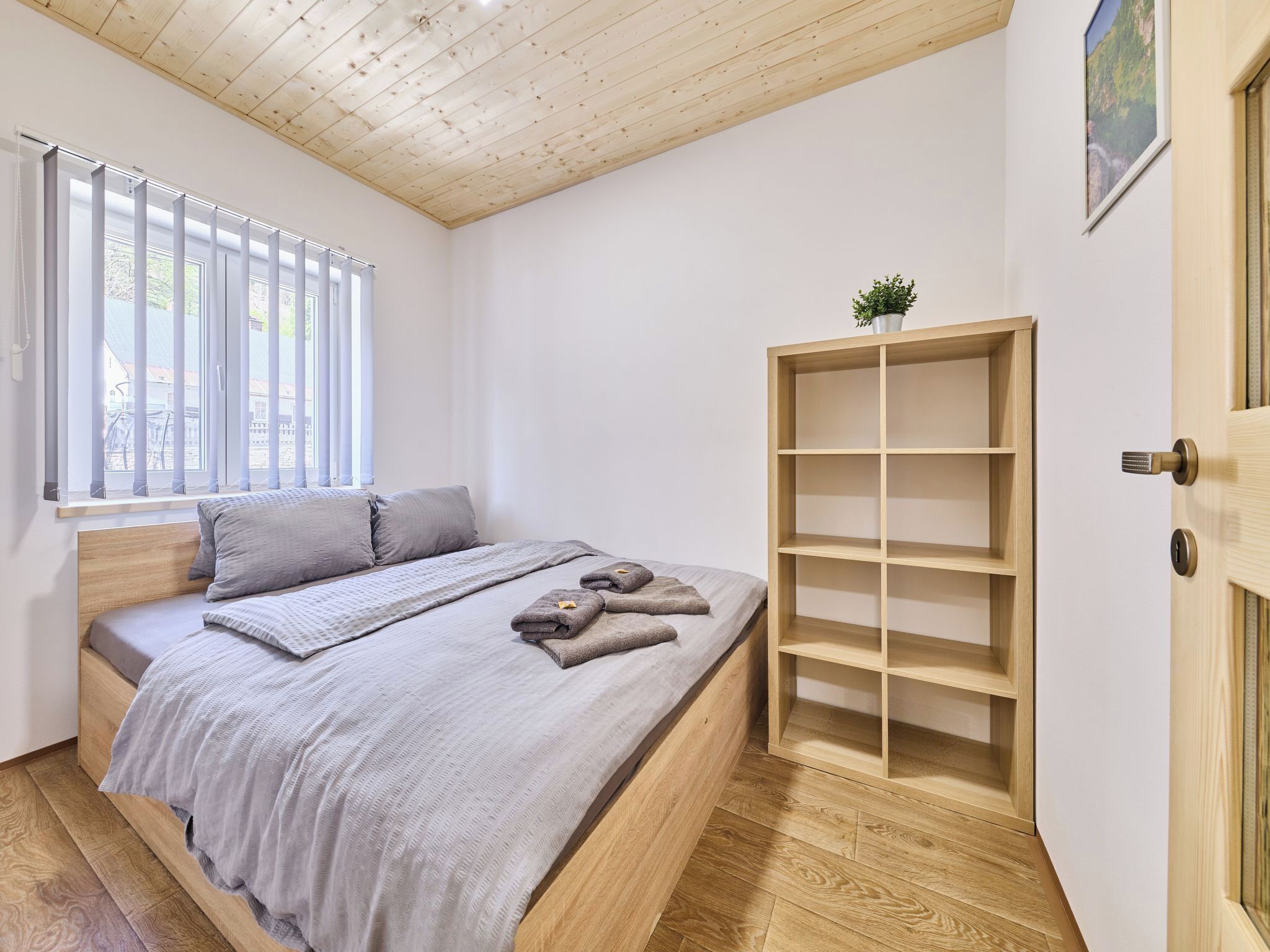 Een slaapkamer met een bed, een raam met zonwering, een houten deur en een inbouwkast.