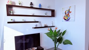 A living room with a flat-screen TV, wooden shelves with decorative items, a potted plant, and a framed artwork on the wall. - Home Sweet Home by the Sea 060 (Durrës)