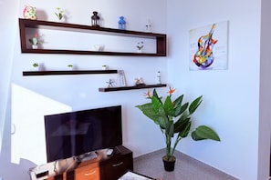 A living room with a flat-screen TV, wooden shelves with decorative items, a potted plant, and a wall art piece. - Home Sweet Home by the Sea 060 (Durrës)