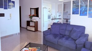 A modern living room with a blue sofa, a coffee table, and a small side table with a vase of flowers. There is a kitchen area visible in the background with white cabinets and a refrigerator. - Home Sweet Home by the Sea 060 (Durrës)