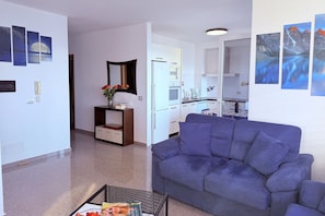 Ein modernes Wohnzimmer mit einem blauen Sofa, einem Couchtisch und einem kleinen Beistelltisch mit Blumen. Im Hintergrund ist eine Küchenzeile mit weißen Schränken und einem Kühlschrank erkennbar.