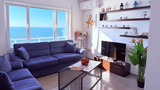 A modern living room with a blue sofa, a coffee table, and a TV. There is a large window with a view of the sea. - Home Sweet Home by the Sea 060 (Durrës)