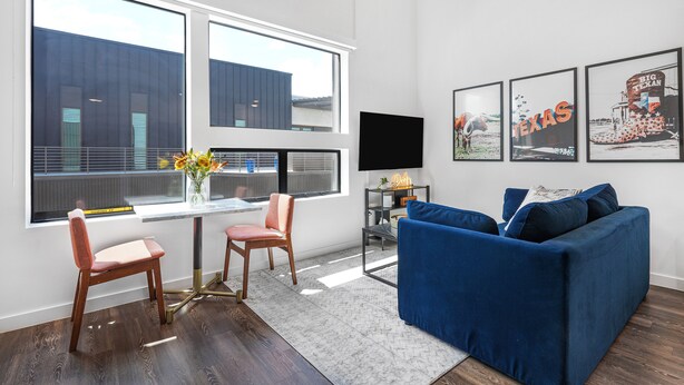 En moderne stue med blå sofa, to stole, sofabord og fjernsyn. Store vinduer giver udsigt til en bygning.