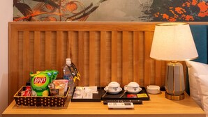 Een hotelkamer met een houten hoofdeinde, een tafel met een mand lekkernijen, een dienblad met thee en kopjes, een lamp en een schilderij aan de muur.