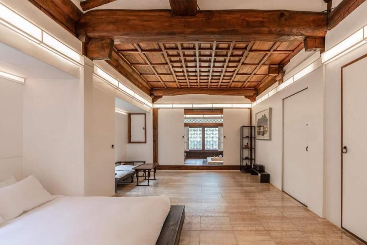 A hotel room with a wooden ceiling, a bed, a desk, and a view of a traditional building through a window.