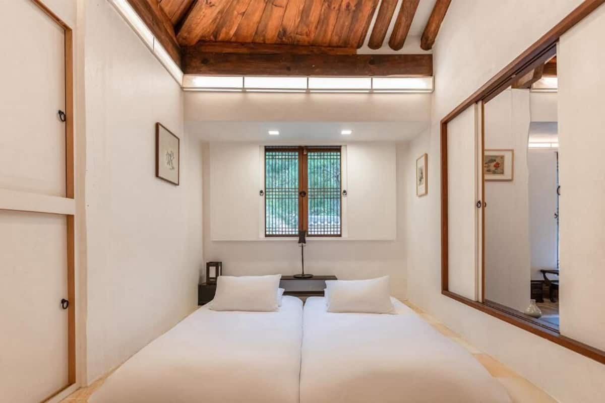 A room with a low ceiling and wooden beams, a bed with white bedding, a window with a view, and a mirror on the wall.