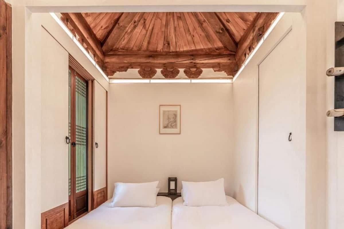 A room with two beds, wooden ceiling beams, a traditional door with a green screen, and a framed picture on the wall.