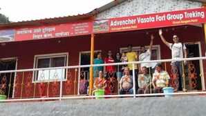 A building with a red facade and a balcony with metal railings. There are people on the balcony and a sign that reads "Advener Vasota Fort Group Trekking". - Jalvishwa agro tourism & River camp Tapola (Satara)