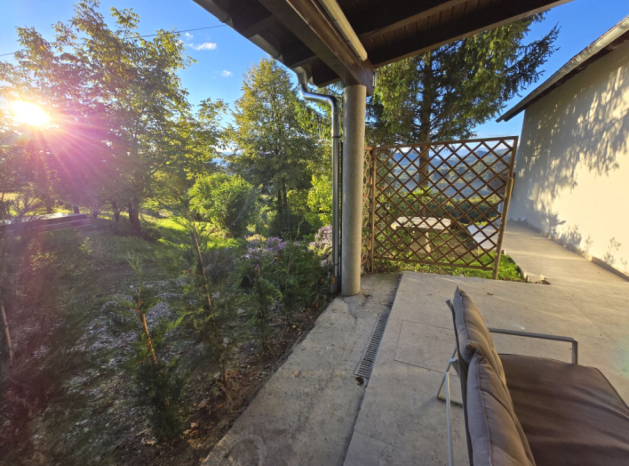 Un patio techado con piso de concreto, cerco de metal y vista a un jardín con plantas y árboles.