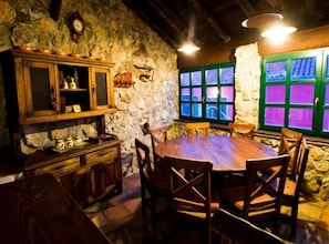 Un interior rústico de piedra con una mesa y sillas de madera, un aparador de madera con estantes y una ventana con contraventanas verdes.