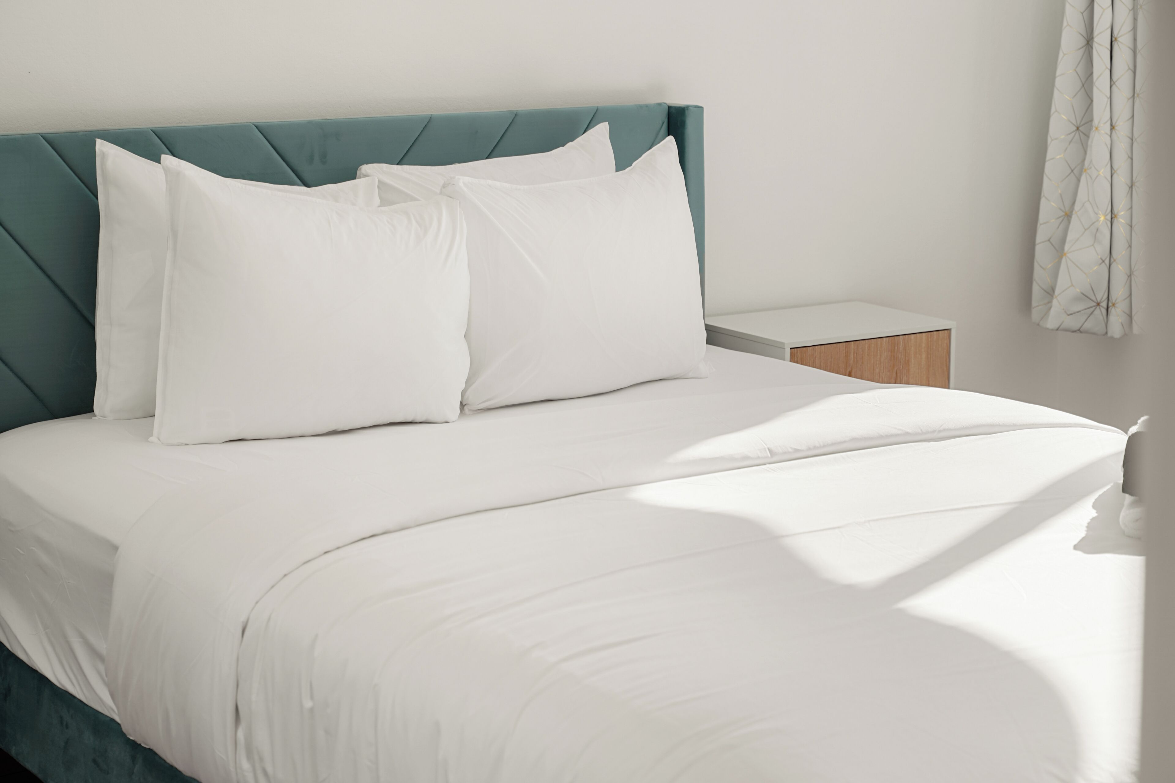 Una cama bien hecha con sábanas y almohadas blancas, un cabecero tapizado en color verde azulado y una mesita de noche con tocador de madera.