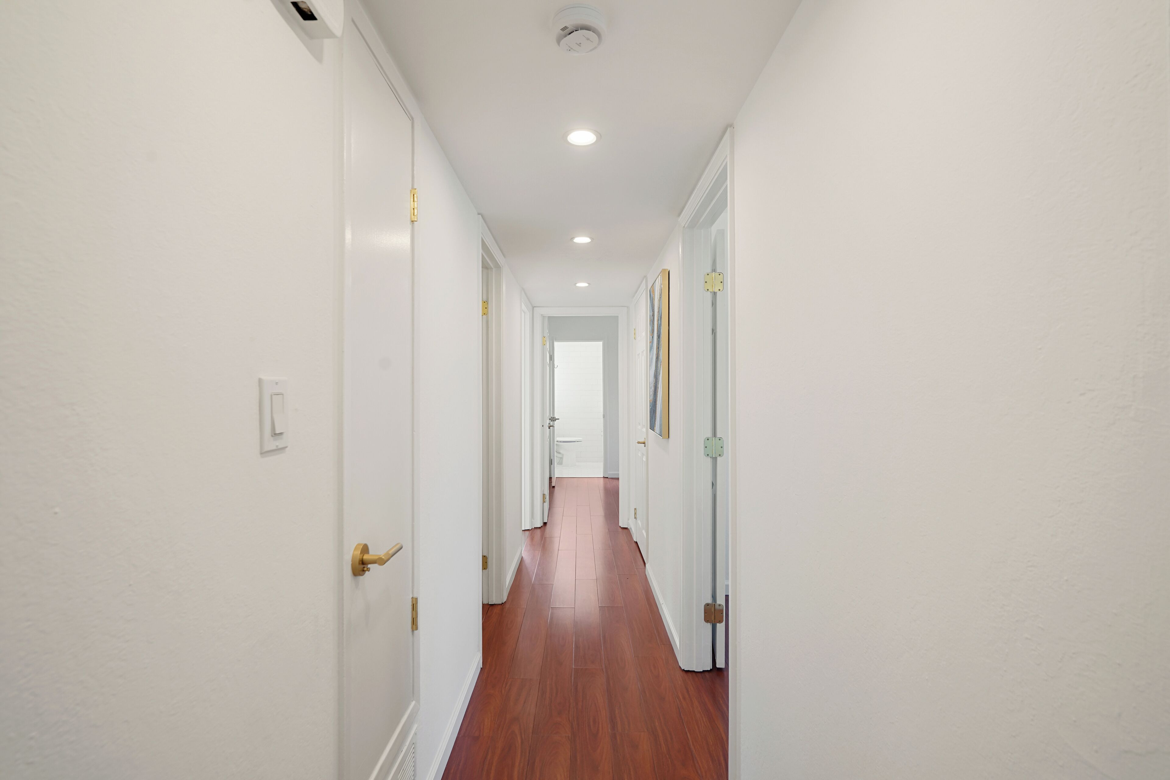 A narrow hallway with white walls and wooden flooring, leading to a door at the end.