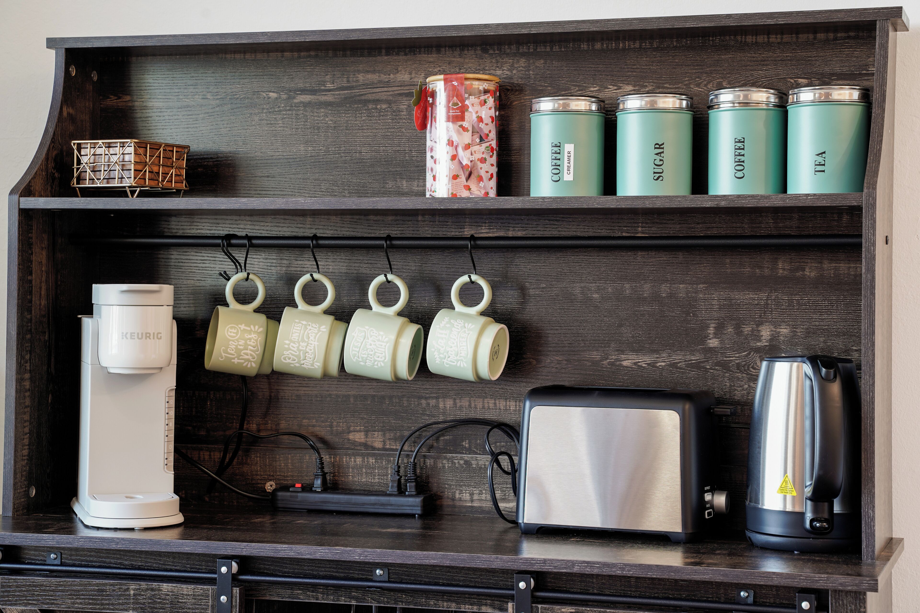 Uma bancada de cozinha com cafeteira, torradeira, chaleira e três recipientes verdes identificados como café, açúcar e chá.