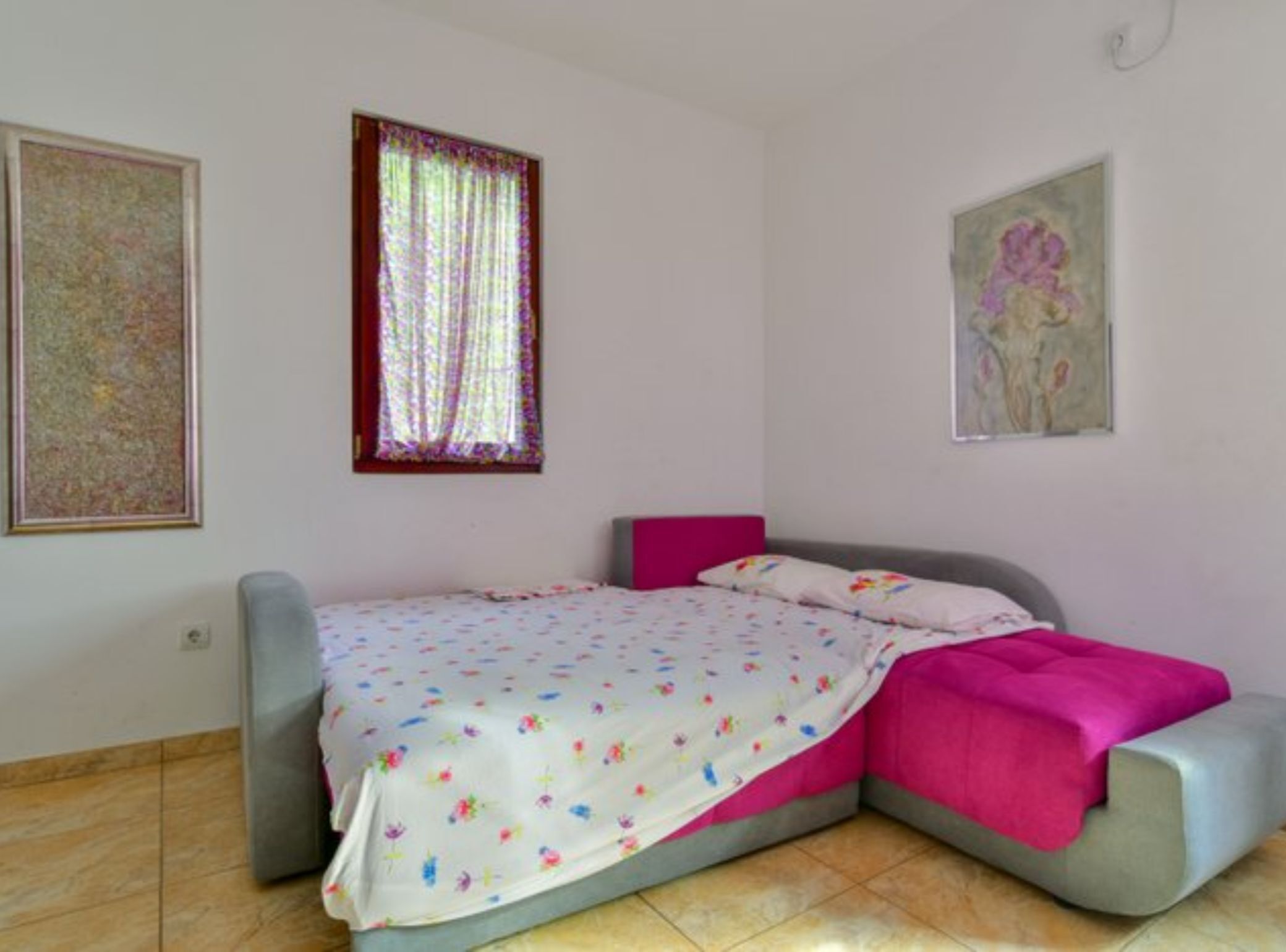 A bedroom with a bed, a window with curtains, and two framed artworks on the wall.