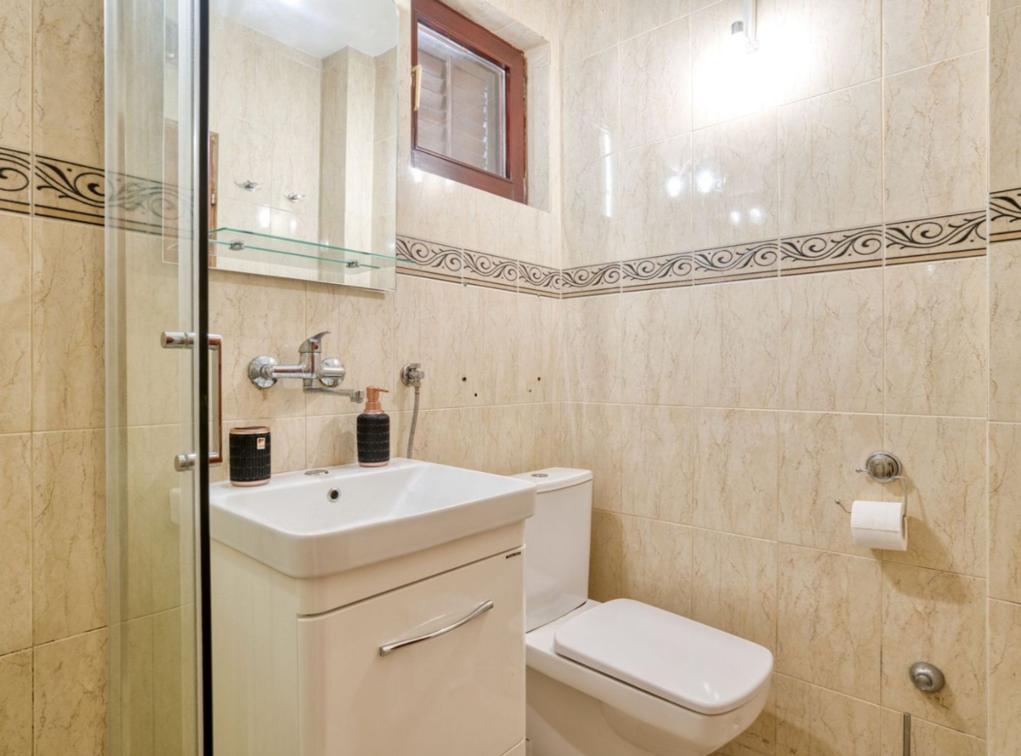 A bathroom with a glass shower enclosure, a white sink with a mirror, a toilet, and a decorative tile border.