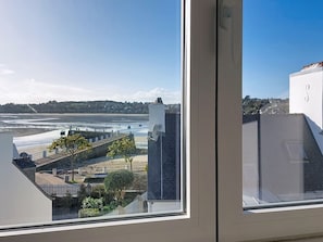 Blick aus einem Fenster auf einen Strand, einen Steg und ein Wohngebiet bei klarem Himmel.