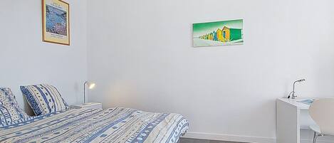 A bedroom with a bed, a desk, a chair, and two wall-mounted paintings.