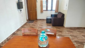 Un coin repas avec une table en bois, deux chaises en bois, une bouteille d’eau, un téléviseur fixé au mur, une porte en bois et une fenêtre avec des rideaux.