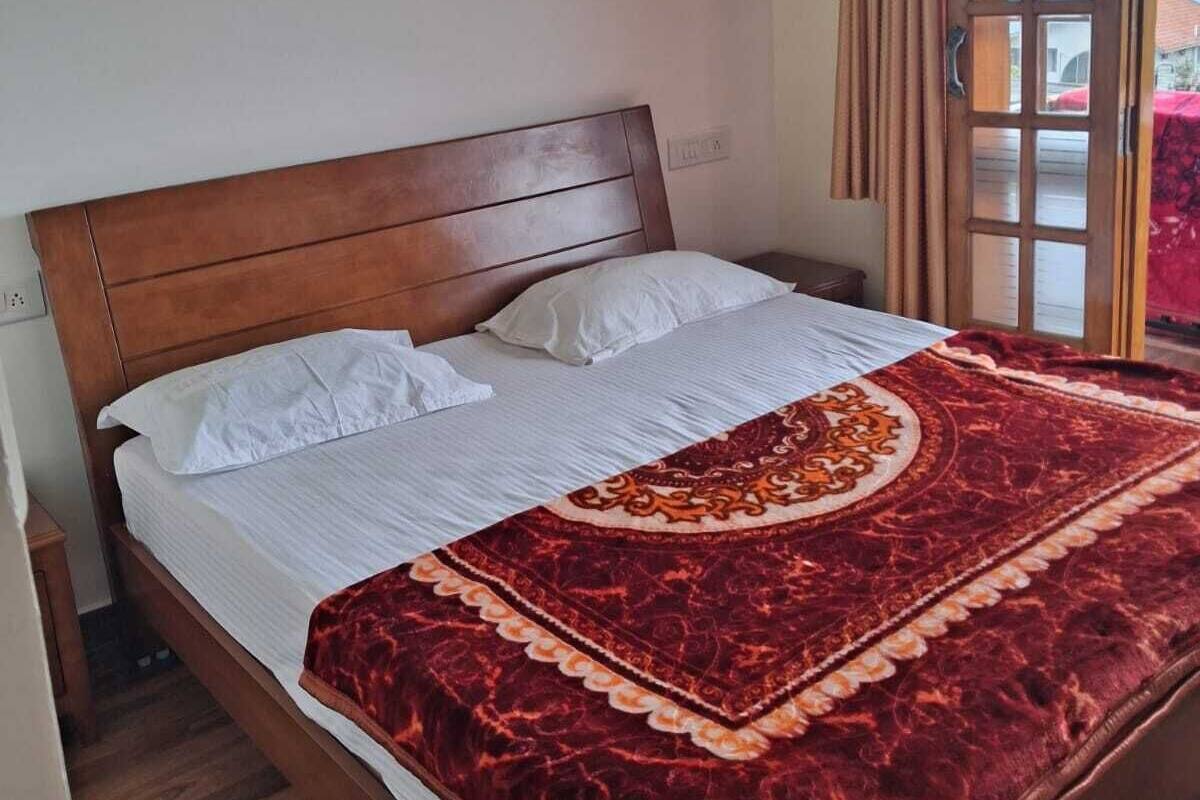 Ein Schlafzimmer mit einem hölzernen Bett, weißer Bettwäsche, einer roten, gemusterten Decke, einer Wandleuchte und einem Fenster mit Vorhängen.