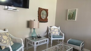 Una sala de estar con muebles de mimbre blanco, una mesa de centro de vidrio, una lámpara y un televisor montado en la pared.