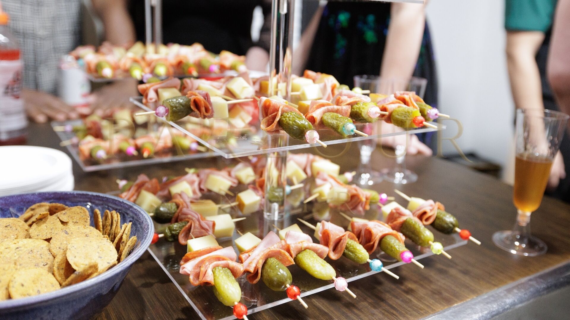 A buffet table with skewers of meat and cheese, a bowl of crackers, and a glass of beer.