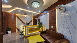 A modern reception area with a marble wall, a wooden reception desk, a chandelier, a fan, and a brown leather sofa. - Hotel Silky Palace (New Delhi)