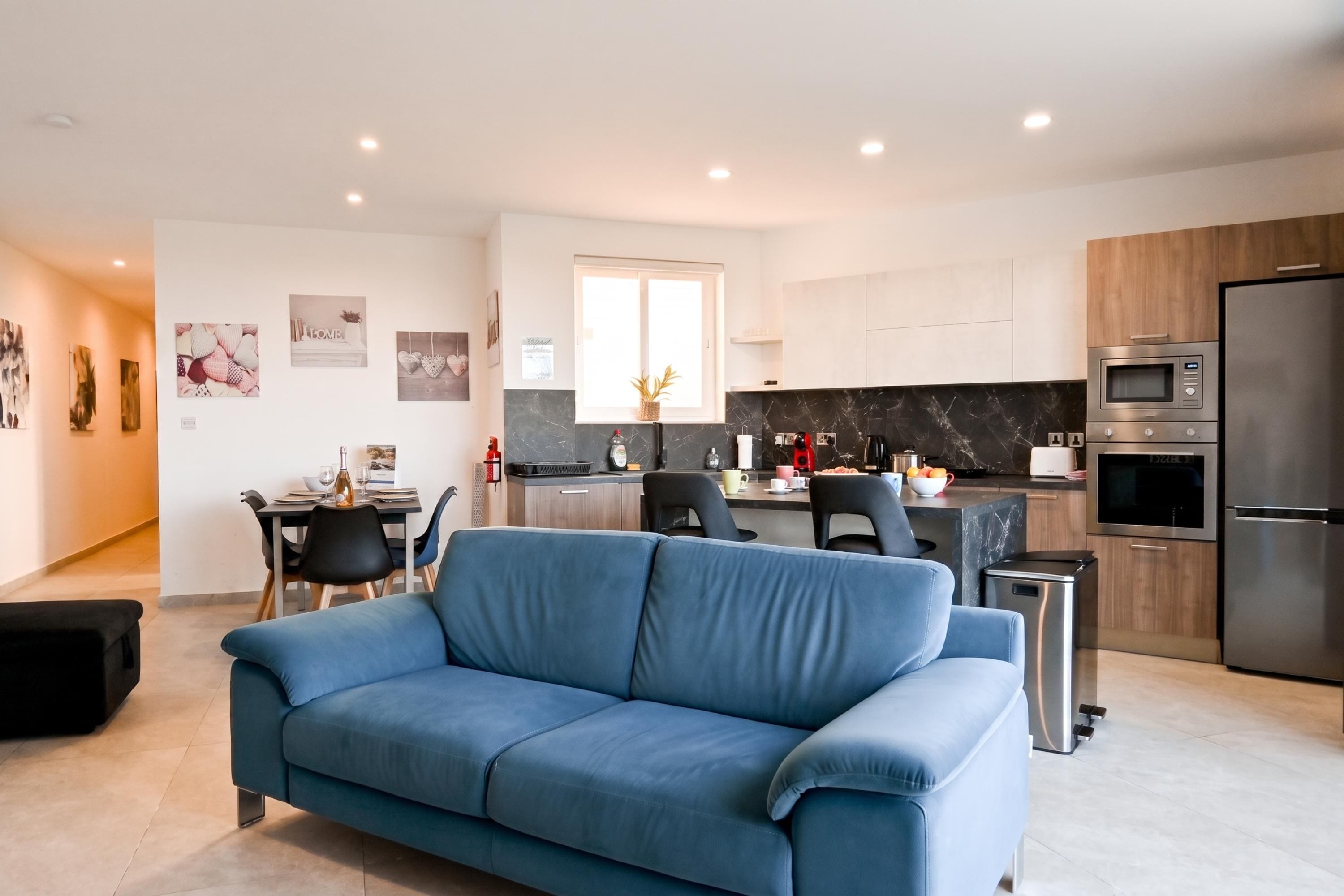 Una cuina moderna amb forn i microones integrats, una zona menjador amb taula i cadires, i un saló amb un sofà blau.