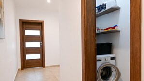 A laundry room with a washing machine, shelves with cleaning supplies, and a wooden door. - Sunny Bay View + shared pool Gozo (Xaghra)