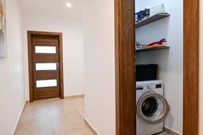 A laundry room with a washing machine, shelves with cleaning supplies, and a wooden door. - Sunny Bay View + shared pool Gozo (Xaghra)