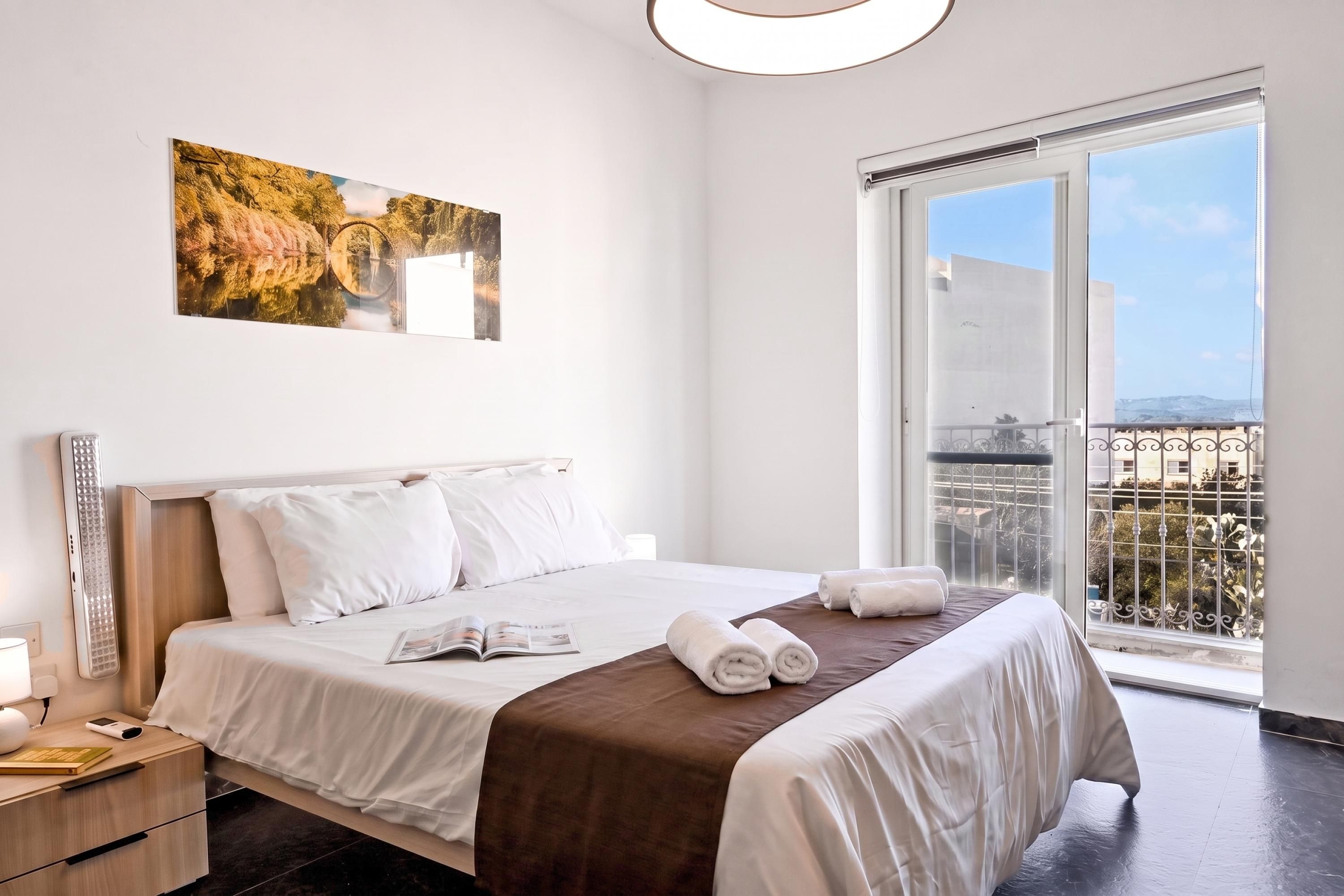 A hotel room with a large bed, a bedside table with a lamp, a window with a view of the city, and a painting on the wall.
