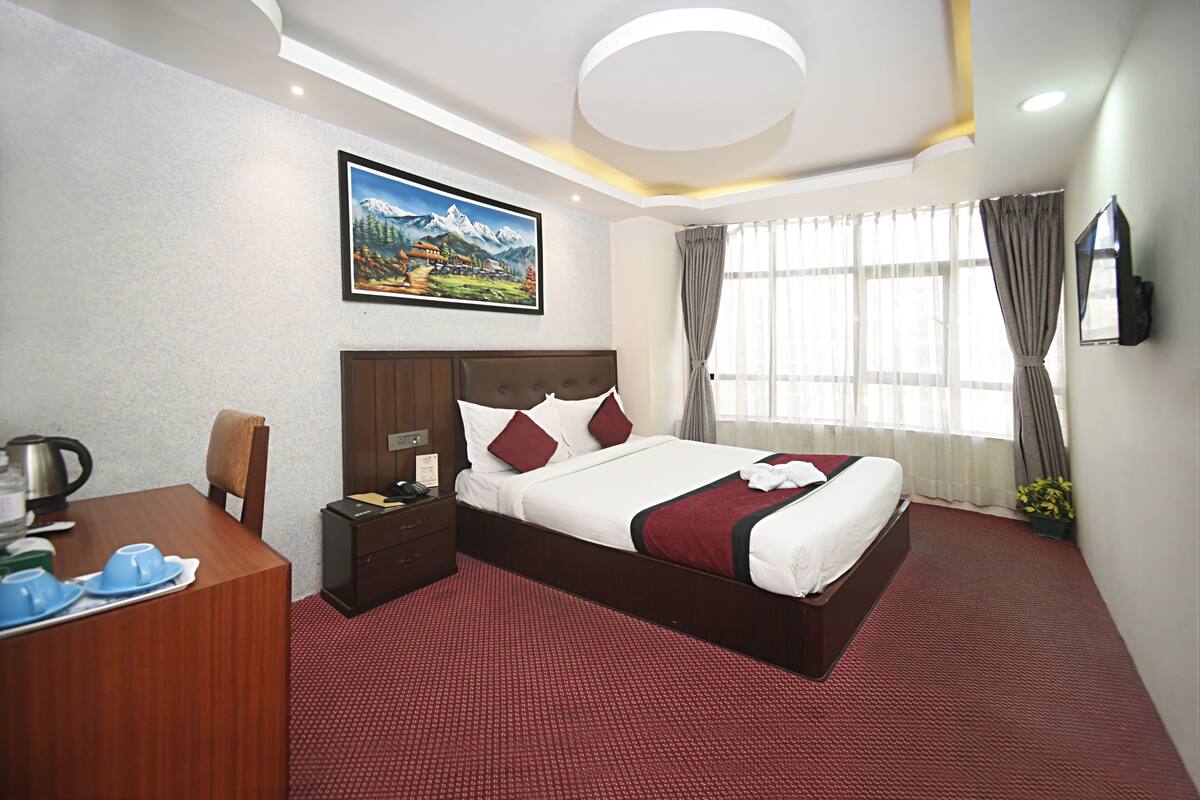 A hotel room with a large bed, a desk with a chair, a painting on the wall, and a window with curtains.