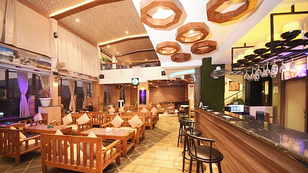 A restaurant with wooden furniture, a bar area, and a high ceiling with wooden light fixtures. - Hotel De Universal (Kathmandu)