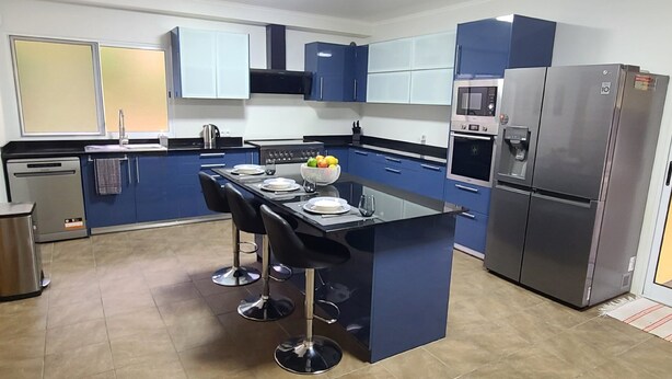 A modern kitchen with blue cabinets, a central island with bar stools, a stainless steel refrigerator, and a dishwasher.