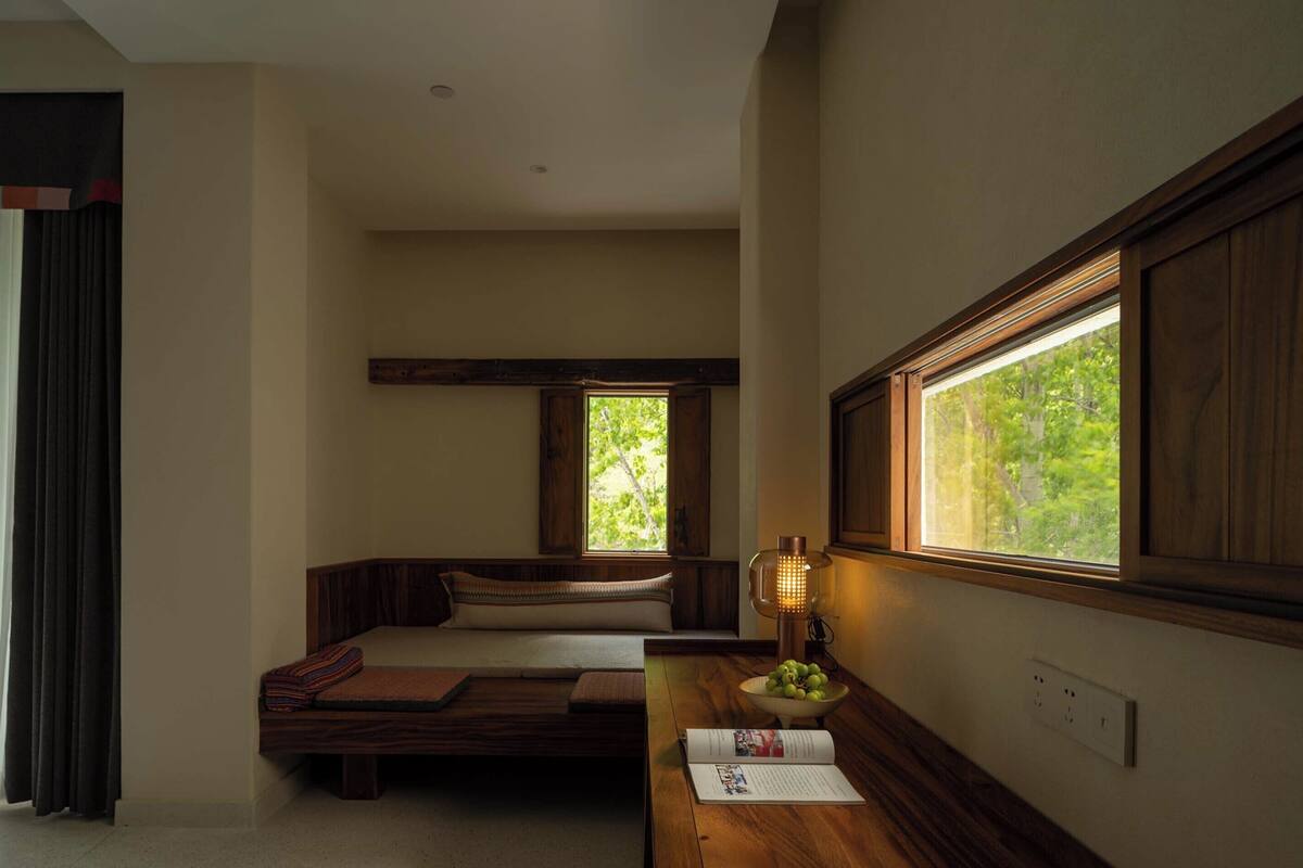 A room with a wooden bench, a table with a lamp, and a window with a view of greenery.