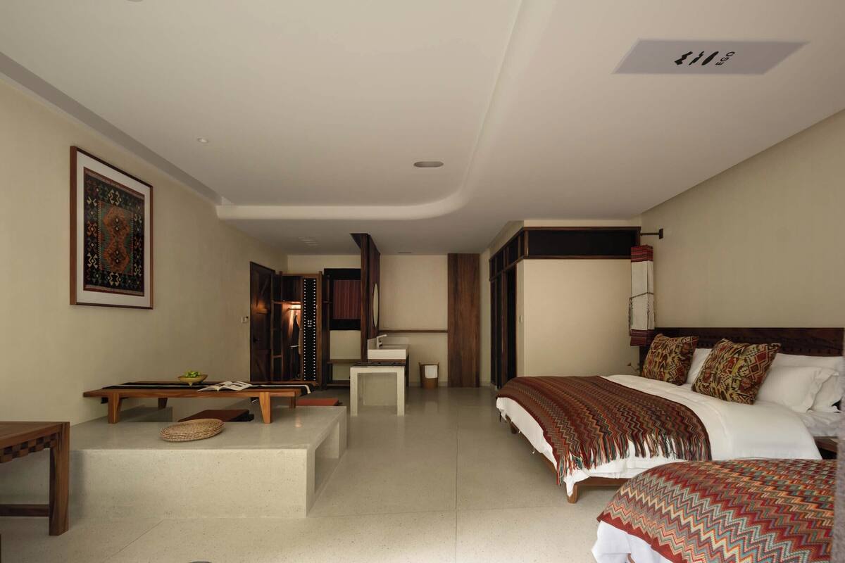 A spacious bedroom with a large bed, a bedside table, a small desk, and a chair. There is a wooden bench with a woven basket and a framed picture on the wall.