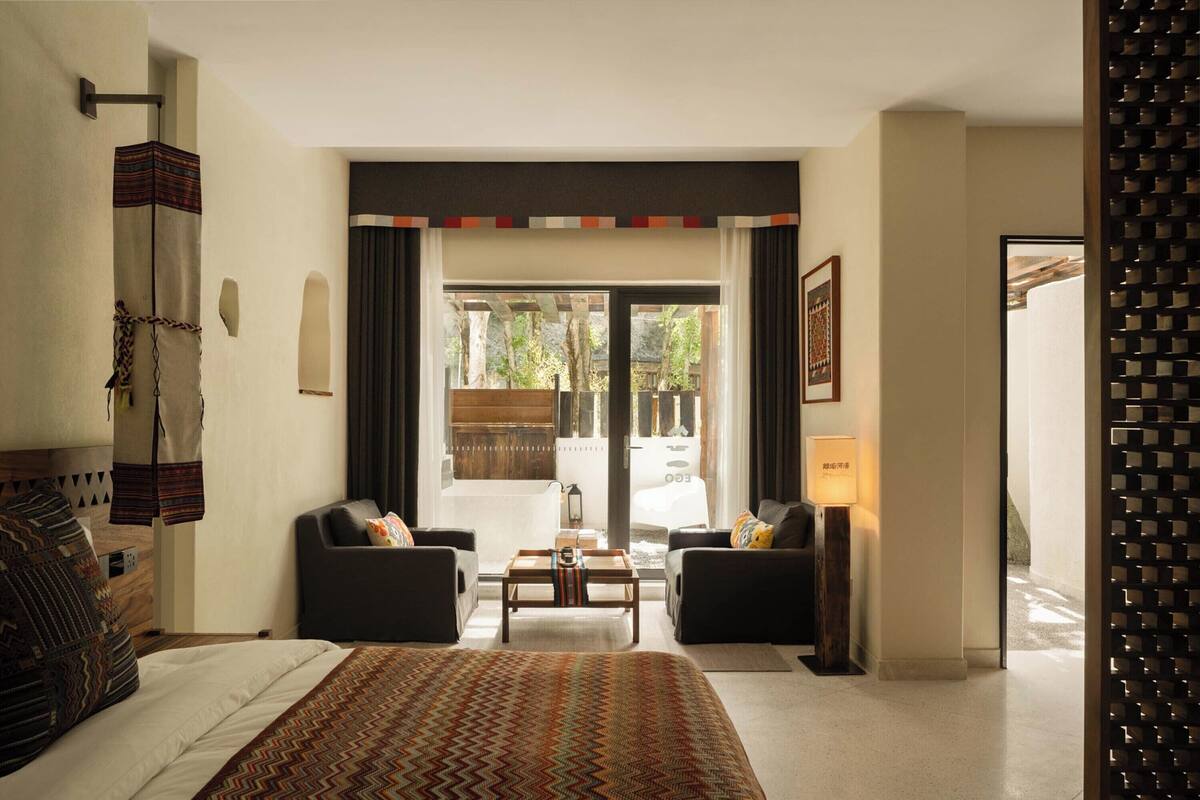 A hotel room with a bed, two armchairs, a coffee table, a lamp, and a view of the outdoors.