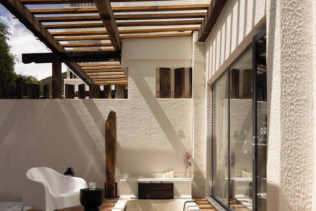 A modern outdoor patio with a wooden pergola, a white chair, a black stool, and a spa tub.