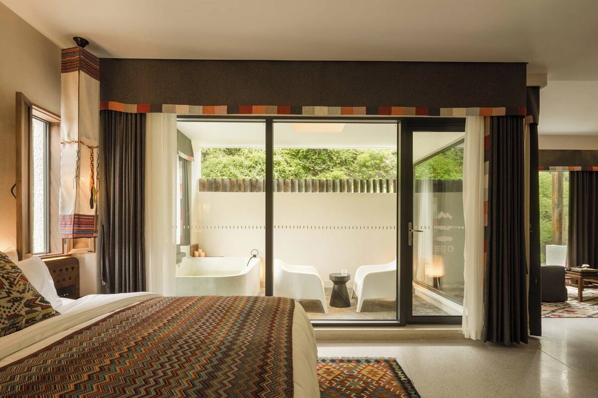 A modern hotel room with a large bed, a glass door leading to a balcony with a view of greenery, a small seating area, and a bathroom with a bathtub.