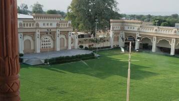 A view of a courtyard with a green lawn, a large tree, and a building with arches in the background.