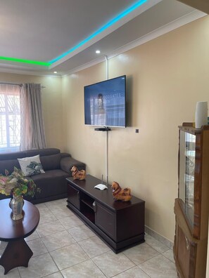 Un salon doté d’un téléviseur à écran plat fixé au mur, d’un meuble de rangement sombre, d’un canapé avec des coussins, d’une petite table sur laquelle est posé un vase, et d’une fenêtre avec des rideaux.