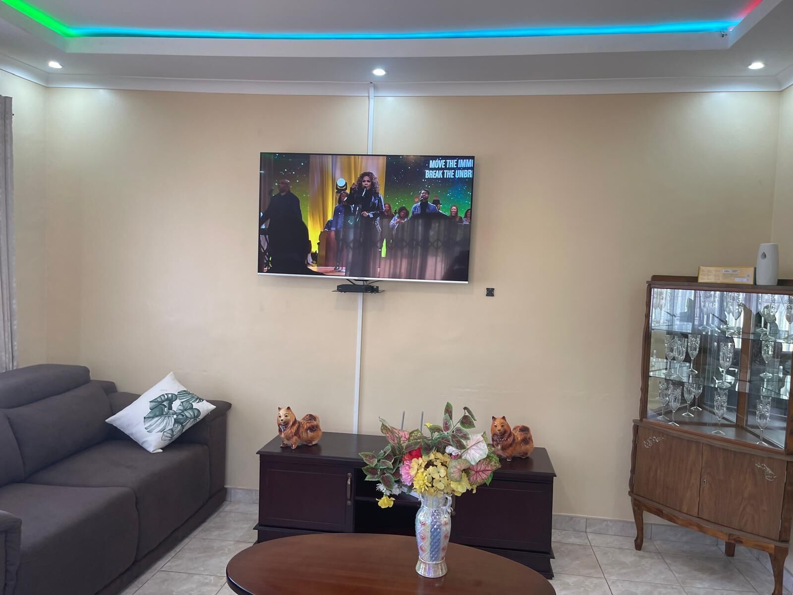 A living room with a flat-screen TV mounted on the wall, a dark brown sofa with a decorative pillow, a wooden cabinet with glass doors, and a round wooden coffee table with a vase of flowers.