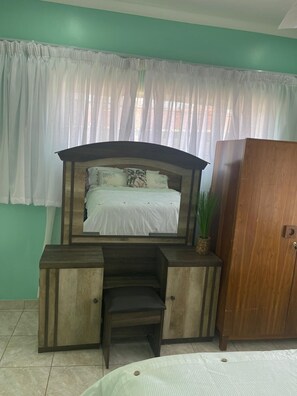 Une chambre à coucher avec un lit, un bureau en bois, une chaise, une armoire et une fenêtre avec des rideaux blancs.