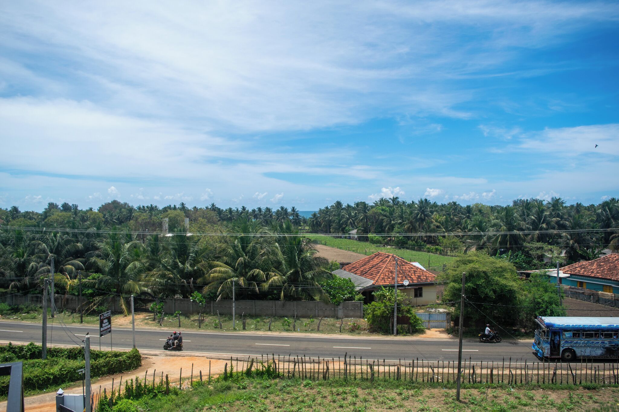 A rural road with a motorbike and a bus, surrounded by greenery and houses with red-tiled roofs.